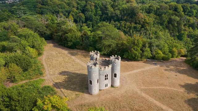 Cinematic drone orbit of historic Blaise Castle in Henbury, Bristol. Elliptical movement reveals the Gothic folly tower surrounded by woodland clearing with visible pathways through dry grass.