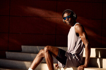 Young athletic man resting on stairs wearing headphones and sunglasses outdoors