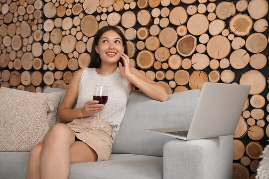 Young Asian woman with wine and laptop having online date on sofa at home - Powered by Adobe