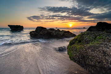 Beautiful sunset seascape with rocks in the ocean, gentle waves, and golden sky reflecting on wet sand, creating a serene tropical beach atmosphere.
