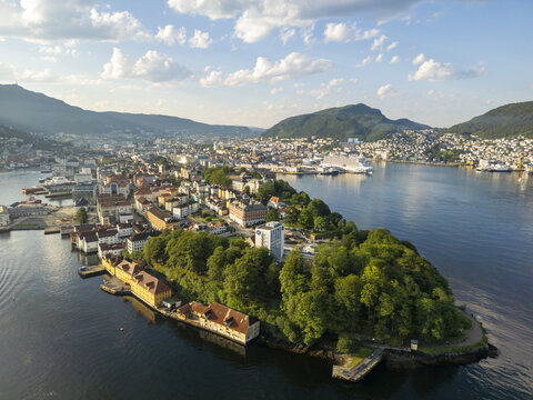 Aerial view of a peninsula with lush greenery and historic buildings jutting into the still, dark waters, framed by distant mountains, Bergen, Vestland, Norway.