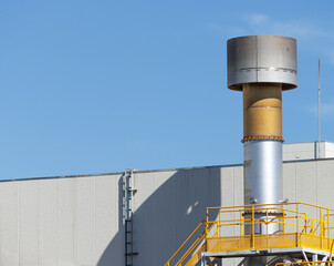 Tall cylindrical grey orange two tone vent pipe stands against clear blue sky. Topped with disk shaped cap and surrounded by orange safety railing walkway, its casts long shadow on adjacent building.
