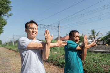 Two men are engaging in stretching exercises in a bright, sunny outdoor area close to train tracks
