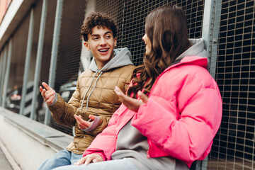 Two teenagers wearing winter jackets are talking and gesturing while sitting in an urban setting