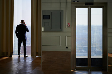 Fototapeta premium Man in suit standing near window with city view, vertical composition, morning light, contemplative posture, office interior, commercial use for business, telecommunication