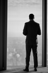 A black-and-white image of a man in a formal suit standing in front of a large window. The man is silhouetted, with his back to the camera, gazing out at the cityscape