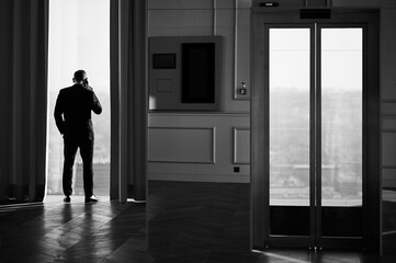 Fototapeta premium A black-and-white photograph captures a man in a formal suit standing near a large window, speaking on the phone. The reflection of the room and window adds depth to the composition