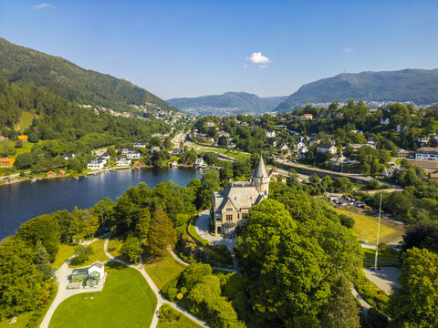 Aerial view of Gamlehaugen royal residence nestled among vibrant green trees beside a tranquil lake, creating a striking contrast with the city beyond, Bergen, Vestland, Norway.