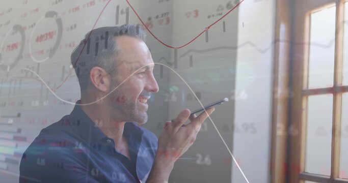 Mature man in blue shirt dictating on smartphone in office by wooden window, with financial charts