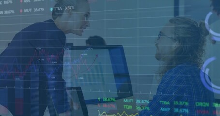 Leaning female in business attire speaking with male colleague at desk, with laptop showing charts