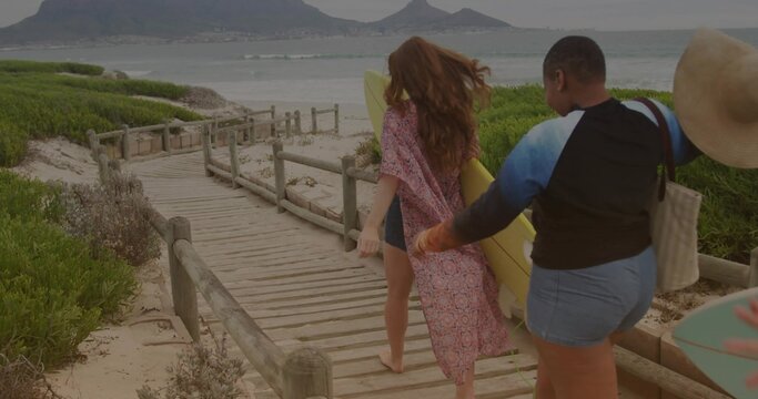 Walking friends in beachwear on beach boardwalk, carrying yellow surfboard, straw hat and tote bag - Powered by Adobe