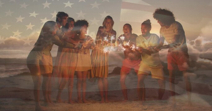 Holding handheld sparklers, eight friends leaning in on sandy beach at sunset, flag overlay shining