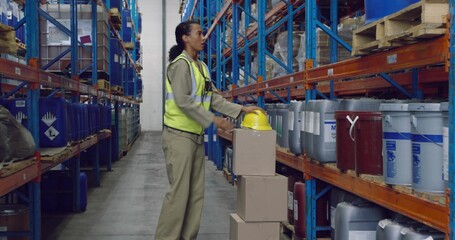 Stacking boxes worker wearing safety vest in storage aisle with hazard drums and yellow helmet