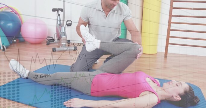 Fototapeta Kneeling trainer bending client right leg on blue mat in rehab studio, with key rehab equipment