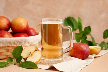 Glass cup of fresh apple cider and wicker basket with fruits on beige background