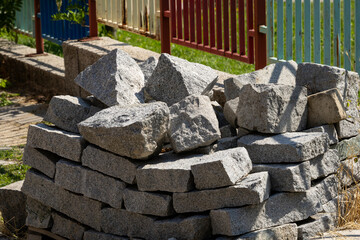 Sunlit rough-cut grey granite blocks stockpiled outdoors for construction or landscaping. Heavy,...
