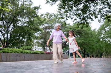Obraz premium Old woman grandmother holding hand with kid girl granddaughter while walking on pavement in park.