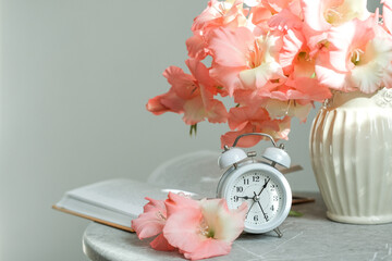 Serene composition featuring white alarm clock, book and vase of pink gladioli on marble table against soft gray background. Feeling of calm and peaceful mornings