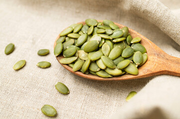 Raw peeled green pumpkin seeds in wooden spoon on natural linen fabric background top view. Food background, Organic healthy tasty pumpkin seeds, Autumn cooking ingredient. Plant-based protein source