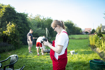 Pregnant woman in red apron standing in garden and holding photo camera, checking pictures on screen. Concept of maternity lifestyle, hobbies during pregnancy, and family leisure. Symbol of expecting