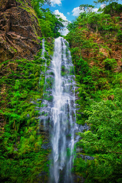 View of a cascading waterfall plunges down a verdant cliff face under a bright blue sky, a striking contrast of textures and colors, Hohoe, Volta Region, Ghana.