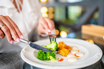 Close-up of hands cutting steamed broccoli on a white plate with grilled tofu and sauces. Cozy dining setting, healthy vegan meal, warm lighting, lifestyle concept