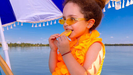 Smiling girl biting lemon slice in yellow swimsuit under umbrella at beach. Concept of playful humor, childhood spontaneity, lifestyle branding, and seasonal creativity.