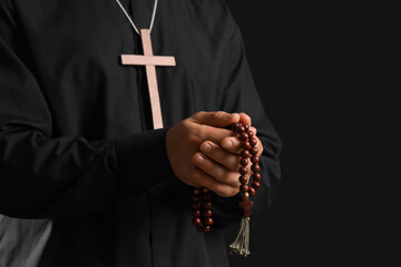 Young priest with wooden cross and beads on black background, closeup