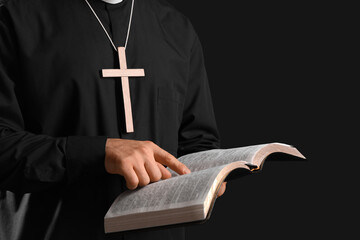Young priest with wooden cross reading Bible on black background, closeup