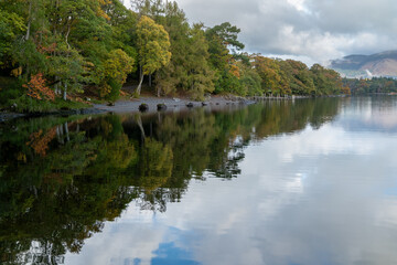Calm lake with autumn trees reflecting on still water under cloudy sky, tranquil shoreline panorama