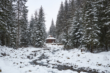 Rustic wooden cabin by a mountain stream in a snowy winter forest.