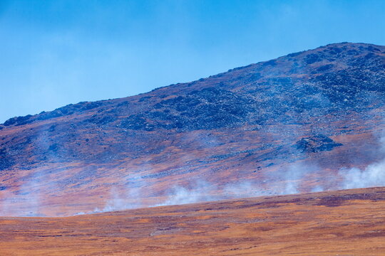 View of a mountain with smoke wafting over the brown and orange landscape under a clear blue sky, Deosai National Park, Gilgit Baltistan, Pakistan.
