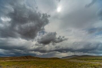 View of a vast, grassy plain meeting low, rolling hills beneath a dramatic, stormy sky with sunlight breaking through, Deosai National Park, Gilgit Baltistan, Pakistan.