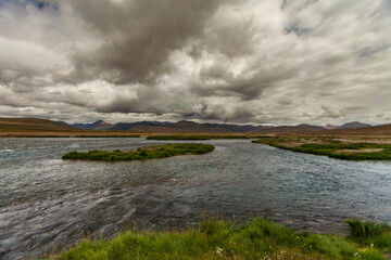 View of a serene river flowing through the vast Deosai plains under a dramatic sky, with mountains in the distance, Deosai National Park, Gilgit Baltistan, Pakistan.