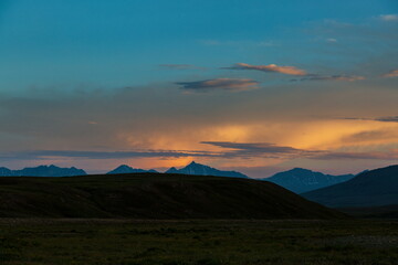 View of rolling hills in shadow beneath a vibrant sky painted with orange and blue hues over distant mountain peaks, Skardu, Gilgit Baltistan, Pakistan.