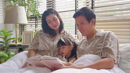Happy Asian family lying together in bed, with mother and daughter cuddling under a warm blanket while father reads a bedtime story from a book. Parents and child bonding through storytelling cozy