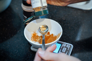 Cook hand pours orange spices into cup on kitchen scale placed on kitchen counter.