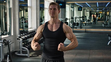 Muscular man in a black tank top flexing his arms and smiling confidently inside a modern gym with weights and exercise equipment. - Powered by Adobe