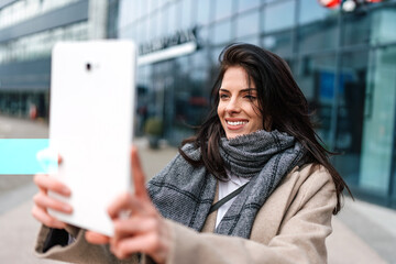 Beautiful fashionable woman standing on city street and using electronic tablet for online...