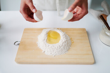 Unknown man breaking egg into mound of flour on wooden cutting board, preparing dough for baking, hands visible holding eggshells above flour and egg mixture