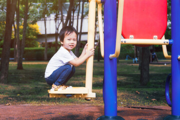 Fototapeta premium Happy Asian boy is playing on the exercise equipment in the outdoor playground, sweet smiling kid aged 3 years old is trying new things. In summer or spring.
