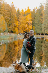 A young Caucasian couple stands by a serene lake surrounded by autumn trees. They hold a bouquet of flowers. Two corgis sit nearby, enjoying the scenery.