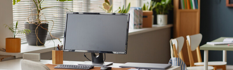 Website header shot of empty classroom desk with computer monitor, keyboard, mouse and notebook positioned near window with potted plants, modern language school workspace prepared for learning