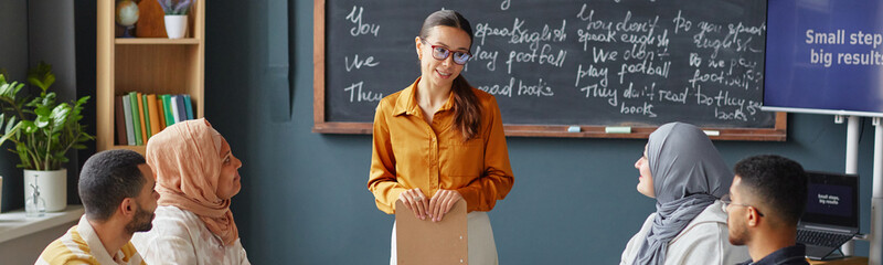 Website header shot of young adult Caucasian woman standing in front of chalkboard teaching diverse...