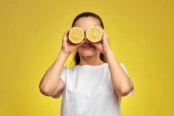 caucasian woman covering eyes with fresh lemon slice on yellow background, vitamin fruit diet