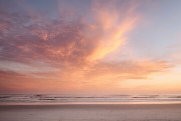 Fototapeta premium Serene beach scene at sunrise, featuring gentle waves and an orangepink hued sky filled with textured clouds. Evokes peace and tranquility, ideal for travel or wellness themes.
