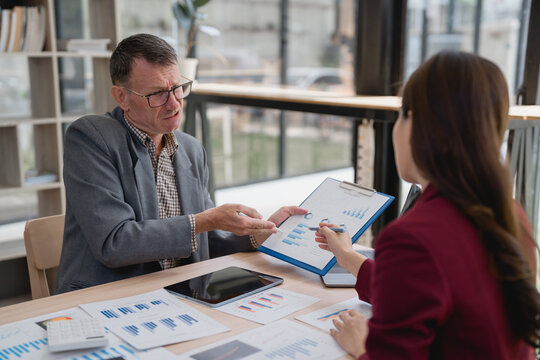 Senior businessman and businesswoman analyzing marketing research charts and graphs, using a clipboard with financial reports and a digital tablet during a corporate meeting in the office - Powered by Adobe