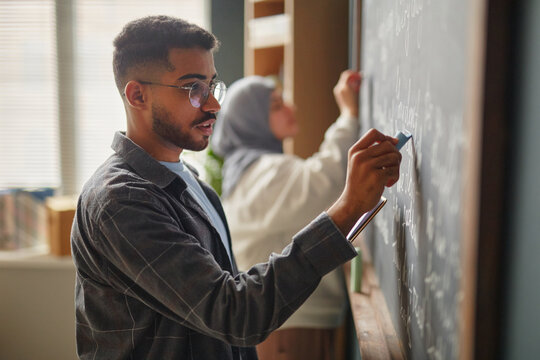 Young adult Black man writing on chalkboard in language school classroom, standing in profile, with young adult Middle Eastern woman in background also writing on board
