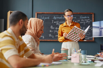 Caucasian young adult woman standing and holding notebook while teaching diverse group of young adult students in language school classroom, students listening and taking notes