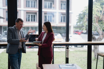 Two businesspeople are discussing a project using a tablet and a smartphone in a modern office near a large window, collaborating and sharing ideas in a professional setting